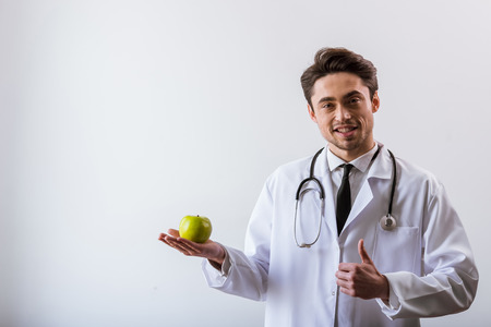 Handsome young doctor in white gown and with stethoscope smiling, looking at camera, holding an apple and showing OK sign, on white backgroundの写真素材