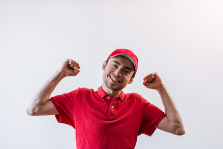 Happy handsome young worker in red t-shirt and cap smiling, looking at camera and keeping hands up, on white backgroundの写真素材