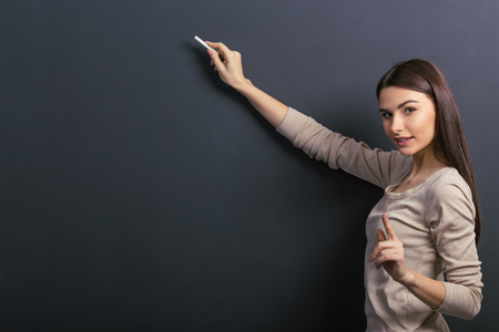 Beautiful young female student is holding a piece of chalk and looking at camera, standing against blackboardの写真素材