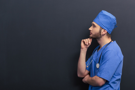 Side view of thoughtful young doctor in blue medical uniform keeping hand on chin and thinking, against blackboardの写真素材