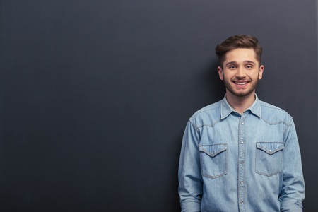 Handsome young student in jeans shirt is looking at camera and smiling, standing against blackboardの写真素材