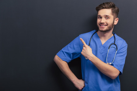 Handsome young doctor in blue medical uniform is pointing away, smiling and looking at camera, standing against blackboardの写真素材