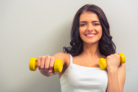 Portrait of attractive young woman holding dumbbells, looking at camera and smiling, against gray background. One hand in focusの写真素材
