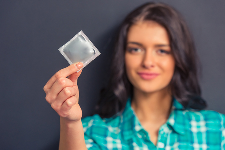 Portrait of attractive girl holding a condom and looking at camera, against dark background. Hand with condom in focusの写真素材