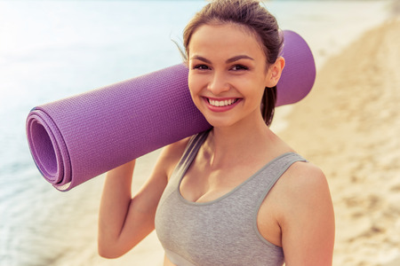 Portrait of beautiful girl in sport clothes holding a yoga mat, looking at camera and smiling, standing on the beach after workoutの写真素材