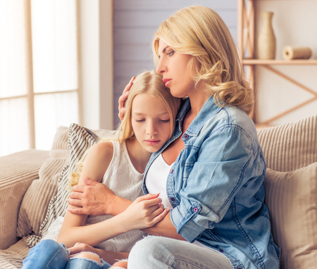 Beautiful blonde woman in jeans shirt and her teenage daughter are hugging while sitting sadly on couch at homeの写真素材