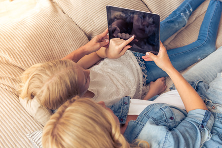 High angle view of beautiful blonde woman in jeans shirt and her teenage daughter using a tablet while lying on couch at homeの写真素材