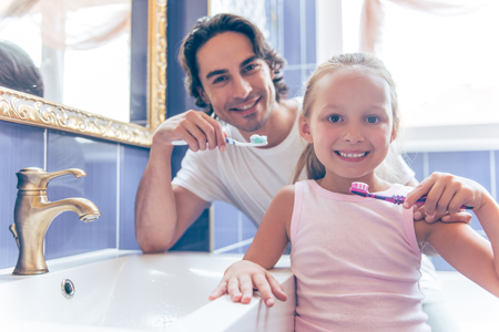 Cute little girl and her handsome father are holding toothbrush with toothpaste, looking at camera and smiling while brushing teeth in bathroom at homeの写真素材