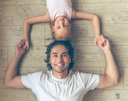 Top view of handsome father and his cute little daughter holding hands, looking at camera and smiling while lying on the floorの写真素材