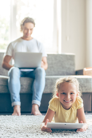 Cute little girl is using a digital tablet, looking at camera and smiling while lying on floor at home, in the background her handsome father is using a laptop, sitting on couchの写真素材