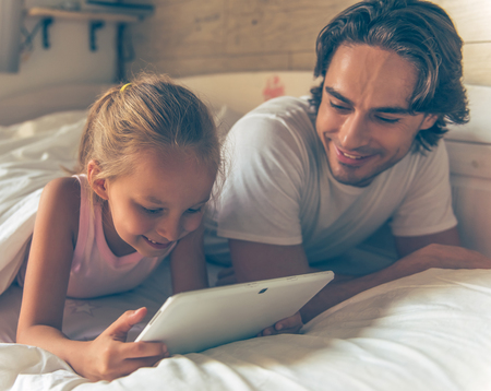 Cute little girl and her handsome father are using a digital tablet and smiling while lying in bed at homeの写真素材