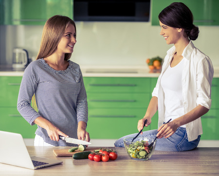 Two beautiful girls are talking and smiling while making salad in the kitchen at homeの写真素材