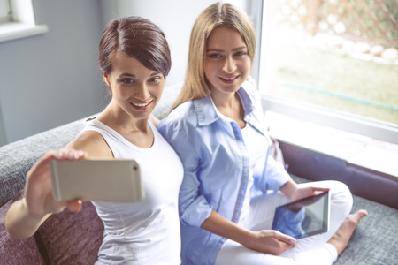 Two beautiful girls are making selfie using a smartphone and smiling while sitting on couch at homeの写真素材