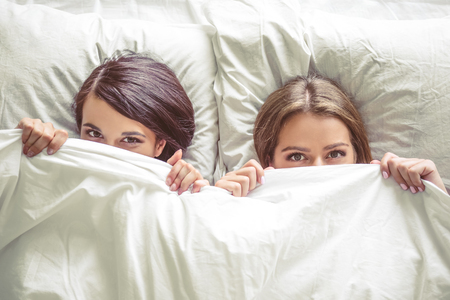 Top view of two beautiful girls hiding under the blanket and looking at camera while lying in bed at homeの写真素材