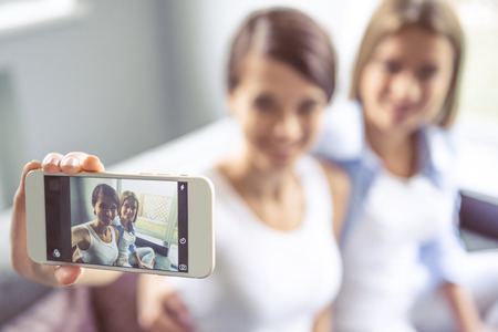 Two beautiful girls are making selfie using a smartphone and smiling while sitting on couch at home. Hand with phone in focusの写真素材
