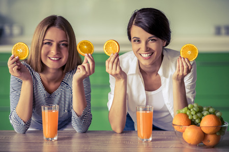 Two beautiful girls are holding slices of orange, looking at camera and smiling while standing in their kitchen at homeの写真素材