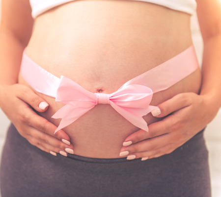 Cropped image of beautiful pregnant woman with pink ribbon on her bare tummy, on white brick wall backgroundの写真素材