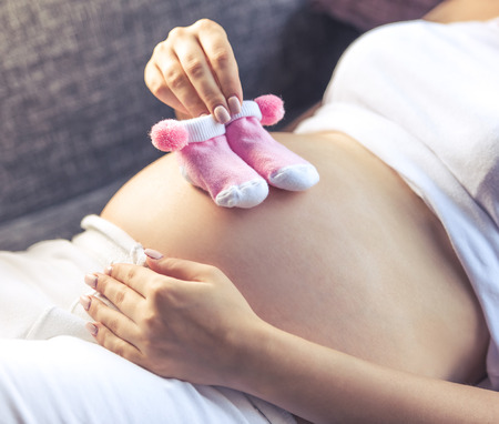 Cropped image of beautiful pregnant woman holding pair of cute baby socks on her bare tummy while lying on the couch at homeの写真素材