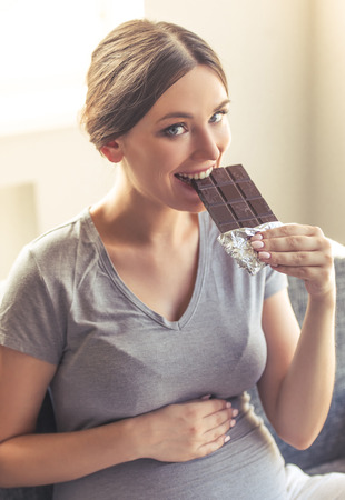 Beautiful pregnant woman is eating chocolate, holding one hand on her tummy, looking at camera and smiling while sitting on the couch at homeの写真素材