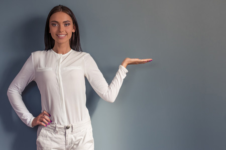Attractive business lady in formal clothes is pointing to the side, looking at camera and smiling, standing on gray backgroundの写真素材