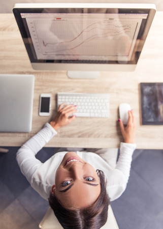 Top view of attractive business lady in stylish clothes using a computer, looking at camera and smiling while working in officeの写真素材