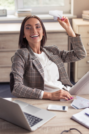 Attractive business lady in stylish formal clothes is holding a paper plane, looking at camera and smiling while sitting at her working place in officeの写真素材