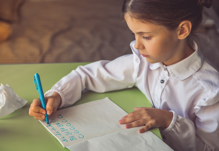 Side view of charming little girl in school uniform doing homework while sitting at the desk at homeの写真素材
