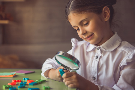 Charming little girl in school uniform is smiling and examining letters using magnifier while doing homework at the desk at homeの写真素材