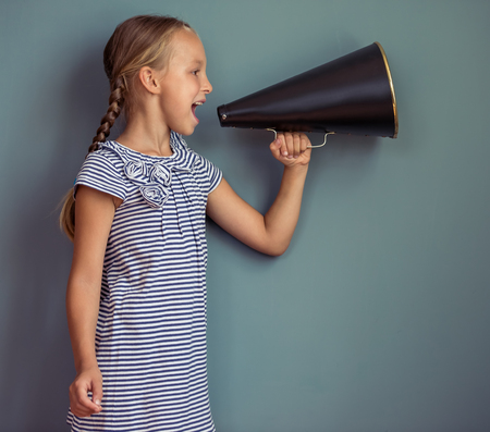 Side view of cute little girl in cute dress screaming into loudspeaker, standing on gray backgroundの写真素材