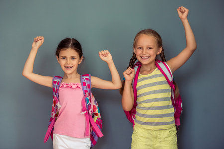 Two cute little girls with school bags are looking at camera and smiling, standing on gray backgroundの写真素材