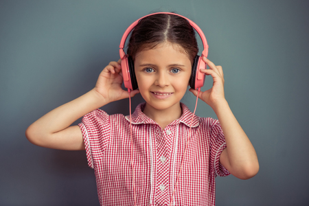 Charming little girl in cute dress and headphones is listening to music, looking at camera and smiling, standing on gray backgroundの写真素材