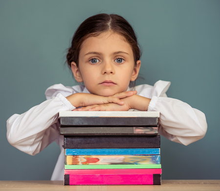 Charming little girl in school uniform is leaning on the pile of books and looking at camera, sitting at the deskの写真素材