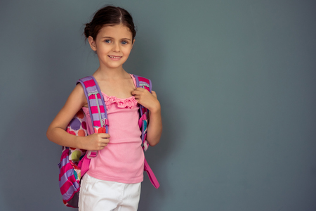 Charming little girl with school bag is looking at camera and smiling, standing on gray backgroundの写真素材