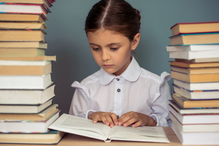 Charming little girl in school uniform is reading a book, sitting between piles of books on gray backgroundの写真素材