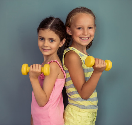 Two cute little girls are holding dumbbells, looking at camera and smiling, standing on gray backgroundの写真素材