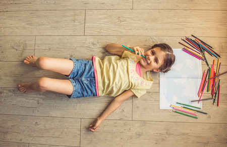 Top view of charming little girl holding a colorful pencil, looking at camera and smiling while drawing, lying on the floor in her room at homeの写真素材