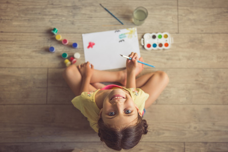 Top view of charming little girl painting, looking at camera and smiling while sitting on the floor in her room at homeの写真素材
