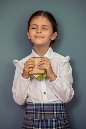 Charming little girl in school uniform is holding a sandwich and smiling, standing with closed eyes on gray backgroundの写真素材
