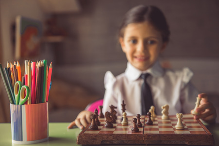 Charming little girl in school uniform and eyeglasses is playing chess, looking at camera and smiling while sitting at the desk at home. Chess board and pencils in focusの写真素材