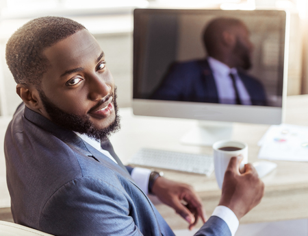Handsome Afro American businessman in classic suit holding a cup of coffee, looking at camera and smiling while working in officeの写真素材