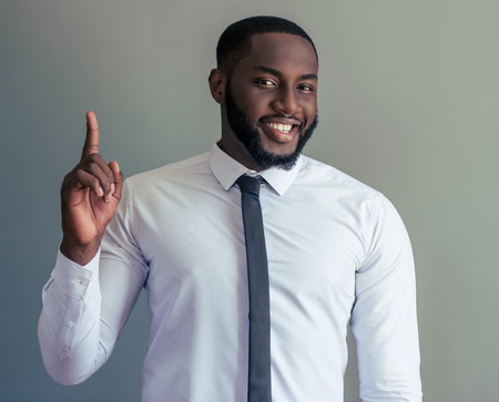 Handsome Afro American businessman in white classic shirt is pointing upward, looking at camera and smiling, on gray backgroundの写真素材