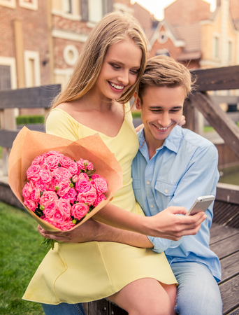 Beautiful young couple is using a smart phone, holding flowers and smiling while sitting outdoorsの写真素材