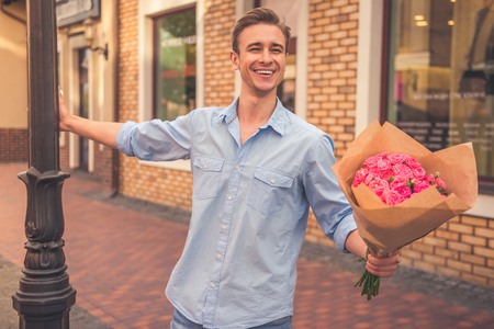 Handsome guy in casual shirt is holding flowers, looking away and smiling while walking outdoors and waiting for his coupleの写真素材