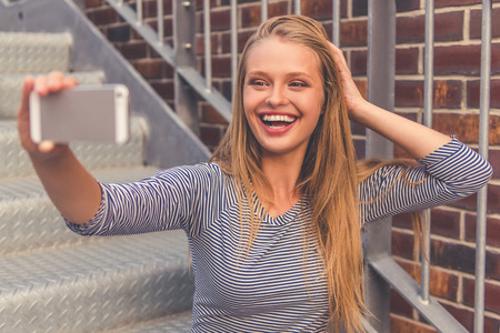 Attractive girl in casual clothes is making selfie using a smart phone and smiling while sitting outdoorsの写真素材