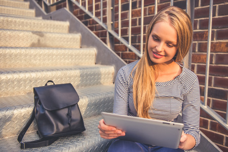 Attractive girl in casual clothes is using a digital tablet and smiling while sitting on stairs outdoorsの写真素材