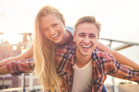 Beautiful young couple is looking at camera and smiling while playing outdoors. Girl is sitting pickabackの写真素材