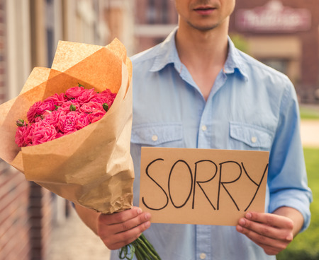 Cropped image of handsome guy in casual shirt holding flowers and piece of paper with word "sorry" while waiting for his coupleの写真素材