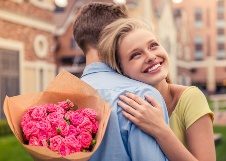 Beautiful young couple on a date. Attractive girl is hugging her boyfriend, holding flowers and smilingの写真素材