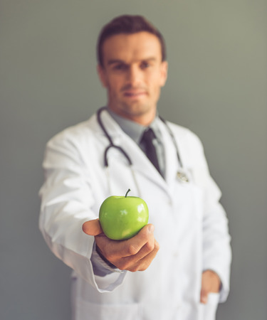 Handsome medical doctor in white coat is holding an apple, on gray background. Hand with apple in focusの写真素材