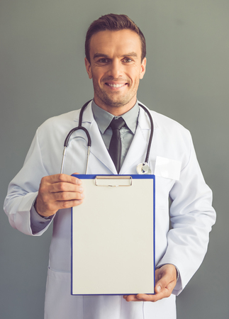 Handsome medical doctor in white coat is holding a folder, looking at camera and smiling while standing on gray backgroundの写真素材